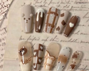 May include: Set of ten long, almond-shaped false nails with various designs. The designs include a bear, hearts, plaid, and a dripping chocolate design. The nails are painted in shades of brown, white, and black.