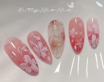 Op de afbeelding: Roze en witte amandelvormige nagels met een kersenbloesemontwerp. De nagels hebben een transparante basis met een roze gradiënt en witte kersenbloesems die erop zijn geschilderd. De nagels zijn versierd met gouden glitter en gouden accenten.