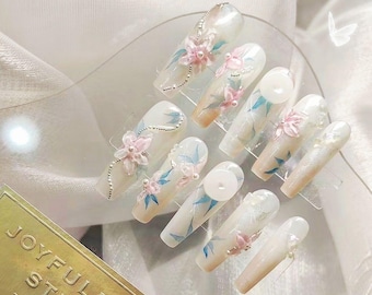 Op de afbeelding: Een set van 10 kunstnagels met een witte basis en een delicaat bloemenontwerp. De nagels hebben roze bloemen, blauwe bladeren en zilveren glitteraccenten. De nagels worden gepresenteerd op een transparante plastic tray met de woorden "Joyful Nail Studio" op een gouden label.