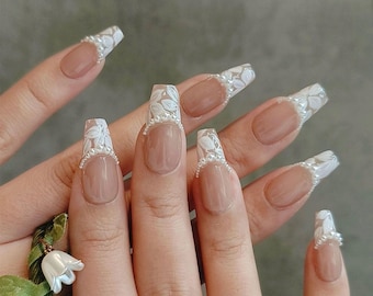 Può includere: Un set di unghie finte color nude con disegni floreali bianchi e dettagli di perle.