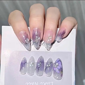 May include: Set of ten false nails with a clear base and a purple and white design. Some nails have small pearl accents and a floral design.