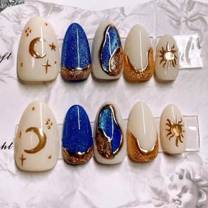 May include: A collection of almond-shaped false nails. Designs feature gold moons, stars, suns, and abstract shapes on a cream and blue glitter base. Gold glitter accents the base of some nails.