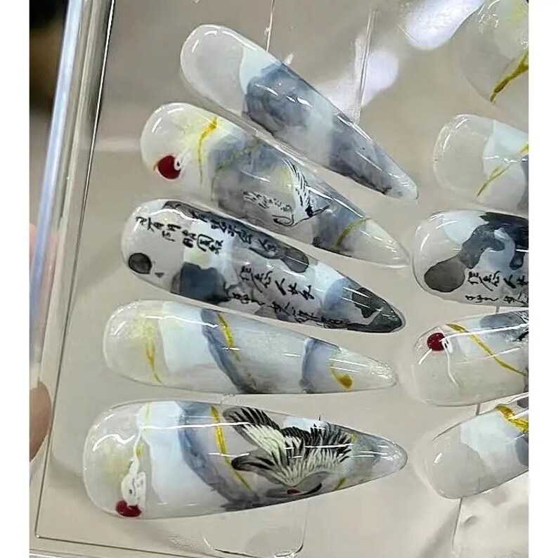 May include: Set of ten false nails with a white, gray, and gold abstract design. The nails feature a watercolor-like effect with calligraphy and a bird design.