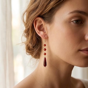 May include: Dangling earrings with a gold-colored hook and a series of red beads. The earrings feature three small round beads and a larger, teardrop-shaped bead at the bottom. The earrings are a simple, elegant design.