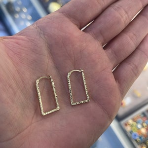 Könnte beinhalten: Ein Paar goldene Creolen mit einem strukturierten, rechteckigen Design. Die Ohrringe werden auf einer Hand gezeigt.