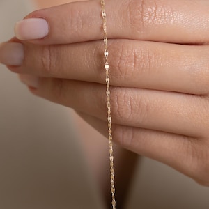 May include: A fine gold necklace featuring a repeating pattern of small, elongated links. The chain is displayed against a neutral backdrop, highlighting its intricate design and gentle gleam. This piece of jewellery is a simple yet elegant accessory.