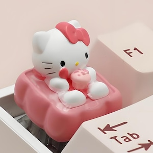 Puede incluir: Una tecla de teclado de Hello Kitty blanca y rosa en un teclado. La tecla presenta una figura de Hello Kitty blanca con un lazo rosa y un asiento rosa. Las teclas del teclado son de color rosa claro.