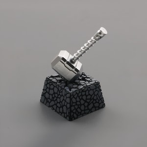May include: A silver metal hammer with a twisted handle sits on a black, textured, square base.