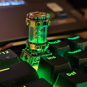 May include: A mechanical-style keyboard keycap featuring a cylindrical, transparent chamber filled with a glowing green spiral structure. The base and top are metallic gray with circular accents, positioned among black keys labeled F12, Ins, and Del.