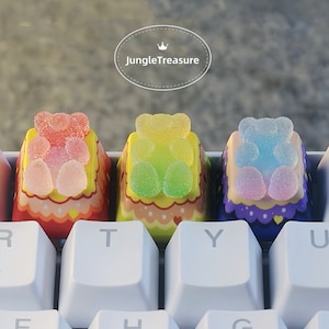 May include: Three colorful gummy bear keycaps with a pastel pink, green, and blue color scheme. The keycaps are decorated with a white border and a heart design. The keycaps are placed on a white keyboard.