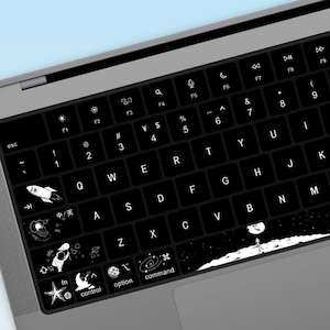 May include: A black laptop keyboard with white keys and a space bar. The keys have various symbols and letters printed on them. The keyboard has a black and white space-themed design with illustrations of planets, stars, a rocket, and an astronaut on the moon.
