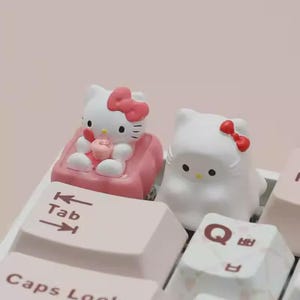 Può includere: Due tasti per tastiera a tema Hello Kitty. Uno è bianco con un fiocco rosa e l'altro è bianco con un fiocco rosso. I tasti sono su una tastiera con tasti rosa e bianchi. Un tasto è seduto su una sedia rosa e tiene in mano un cupcake.
