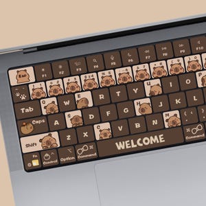 Könnte beinhalten: Eine braune Laptop-Tastatur mit Comic-Capybara-Illustrationen auf jeder Taste. Die Leertaste sagt "WELCOME".
