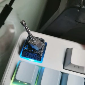 Puede incluir: Un tapón de tecla de teclado plateado y negro con un diseño de martillo en miniatura. El tapón de tecla está iluminado con una luz azul.