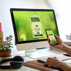 Może przedstawiać: Ekran komputera i telefon kom&oacute;rkowy wyświetlają reklamę "Rubbel K&auml;rtchen" w kolorze zielonym i złotym z motywem koniczyny. Tekst na reklamie brzmi "Gl&uuml;ck ist das einzige, das sich verdoppelt, wenn man es teilt."