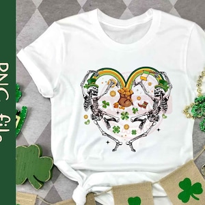 May include: White t-shirt with a graphic of two skeletons dancing in a heart shape with a rainbow and shamrocks. The skeletons are wearing green hats and holding gold coins.