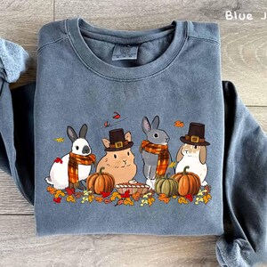 May include: Blue jean colored sweatshirt featuring a fall-themed graphic. The graphic includes four cartoon rabbits and a hamster, wearing hats and scarves, surrounded by pumpkins and autumn leaves. The text "PNG FILE" is on the left side of the image.