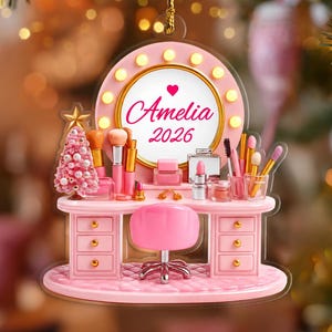 May include: A pink vanity ornament featuring a round mirror with lights, a pink chair, and makeup brushes. The mirror displays the name Amelia and the year 2026 in pink script. A small pink Christmas tree stands on the left side of the desk.