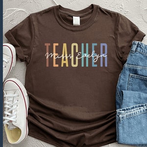 Puede incluir: Una camiseta marr&oacute;n con la palabra "TEACHER" en colores arco&iacute;ris y el texto "Miss Evelyn" en letra blanca.
