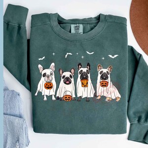 Pu&ograve; includere: Una felpa verde scuro con una grafica di quattro Bulldog francesi vestiti da fantasmi per Halloween. Ogni cane tiene una piccola zucca. Il testo "COMFORT COLORS" &egrave; visibile sull'etichetta.