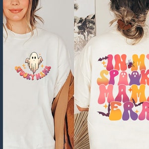 Op de afbeelding: Twee witte T-shirts met een Halloween-thema. De voorkant van het ene shirt heeft een spook met de tekst "Spooky Mama" in regenboogkleuren. De achterkant van het andere shirt heeft de tekst "In My Spooky Mama Era" in regenboogkleuren met een spin, een spinnenweb en vleermuizen.