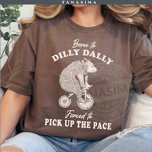 May include: A brown t-shirt featuring a white graphic of a bear on a bicycle, with the text "Born to Dilly Dally, Forced to Pick Up the Pace." The shirt has a casual style and a relaxed fit.