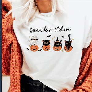May include: White t-shirt with the text "Spooky Vibes" and a graphic of four black cats dressed in Halloween costumes sitting on pumpkins.