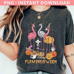 May include: A black t-shirt with a graphic design of pink flamingos dressed in Halloween costumes. The flamingos are holding pumpkins and candy. The text "Flamingoween" is printed below the flamingos.