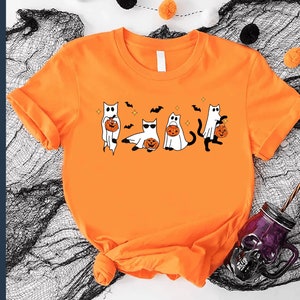 May include: Orange t-shirt featuring a Halloween-themed graphic. The design includes cartoon cats dressed as ghosts, holding jack-o'-lanterns, with bats and stars. The shirt is made of a soft material and is perfect for the spooky season.