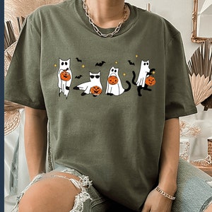 May include: Olive green t-shirt featuring a Halloween-themed graphic. The design includes four cartoon cats dressed as ghosts, each holding a jack-o'-lantern. Bats and stars are also included in the design. The shirt is a PNG file.