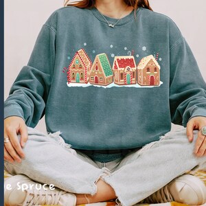 May include: A teal blue sweatshirt with a graphic of five gingerbread houses with red, white, and green icing. The houses are decorated with candy canes, sprinkles, and stars.