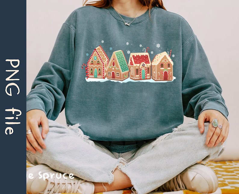 May include: A teal blue sweatshirt with a graphic of five gingerbread houses with red, white, and green icing. The houses are decorated with candy canes, sprinkles, and stars.