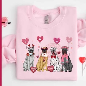 Puede incluir: Sudadera rosa claro con un diseño de cuatro perros con accesorios de San Valentín. Cada perro sostiene un globo en forma de corazón con la palabra "LOVE". Llevan gafas de sol, pajaritas y coronas de flores, ideal para San Valentín.