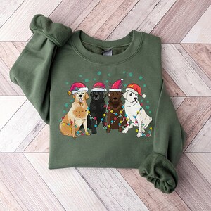 Puede incluir: Una sudadera verde con un dise&ntilde;o navide&ntilde;o que presenta cuatro perros con gorros de Pap&aacute; Noel y rodeados de luces navide&ntilde;as de colores.