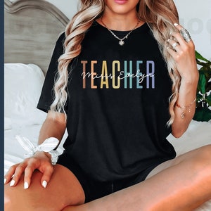Puede incluir: Una camiseta negra con el texto "TEACHER" en colores arco&iacute;ris y el texto "Miss Evelyn" en blanco debajo.