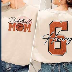 May include: A beige sweatshirt with the text "Basketball Mom" in a red, white, and black design. The sweatshirt also features a basketball graphic. The back of the sweatshirt has a large orange number 6 with the text "Henry" in black.