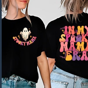 Op de afbeelding: Een zwart T-shirt met een grafisch ontwerp van een spook en de tekst "Spooky Mama" in regenboogkleuren. De achterkant van het shirt heeft de tekst "In My Spooky Mama Era" in regenboogkleuren met een spin, vleermuizen en een spinnenweb.