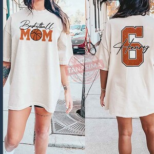 May include: Two white t-shirts, one with the text "Basketball Mom" and a basketball graphic, the other with the number "6" and the name "Henry" in a script font.