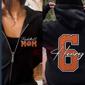May include: Two black hoodies with different designs. One hoodie has the text "Basketball Mom" with a basketball graphic. The other hoodie has the number "6" with the name "Henry" written underneath.