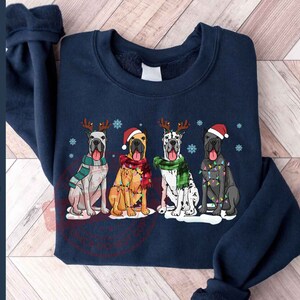 May include: A navy blue sweatshirt with a Christmas-themed design featuring four Great Danes wearing Santa hats and Christmas lights. The dogs are standing in a row, with their tails wagging.