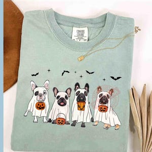 Pu&ograve; includere: Una maglietta verde chiaro con un design di quattro bulldog francesi vestiti da fantasmi che tengono zucche. Il testo "Comfort Colors" &egrave; sulla maglietta.