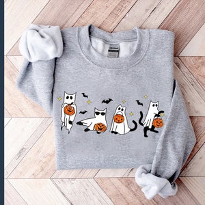 May include: A light gray sweatshirt featuring a Halloween-themed design. The design includes cartoon cats dressed as ghosts, holding jack-o'-lanterns, with bats and stars. The sweatshirt has long sleeves and a crew neck.