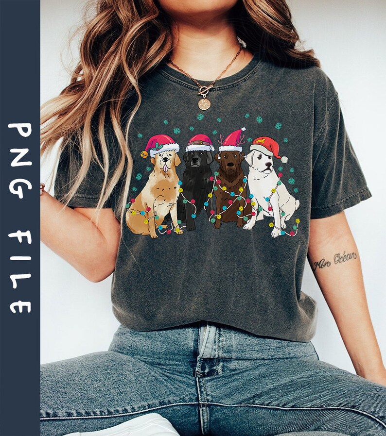 Puede incluir: Una camiseta gris oscuro con un gr&aacute;fico de cuatro perros con gorros de Pap&aacute; Noel y rodeados de luces navide&ntilde;as. Los perros son un golden retriever, un labrador negro, un labrador chocolate y un perro blanco.