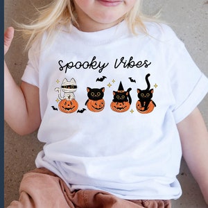 May include: A white t-shirt with the text "Spooky Vibes" and a graphic of four black cats sitting in pumpkins. The cats are wearing different Halloween costumes, including a mummy, a witch's hat, and a regular black cat.