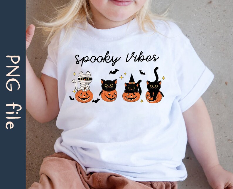 May include: A white t-shirt with the text "Spooky Vibes" and a graphic of four black cats sitting in pumpkins. The cats are wearing different Halloween costumes, including a mummy, a witch's hat, and a regular black cat.