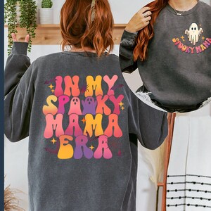 Op de afbeelding: Twee sweatshirts met een Halloween-thema. Een sweatshirt is zwart met de tekst "In My Spooky Mama Era" in regenboogkleuren. De andere sweatshirt is zwart met een witte geest en de tekst "Spooky Mama".