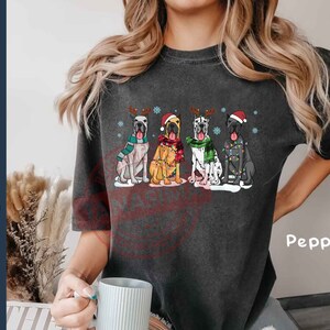 May include: A dark gray t-shirt with a graphic of four Great Danes wearing Santa hats and reindeer antlers. The dogs are wearing festive scarves and have Christmas lights around them. The text "Pepper" is visible in the bottom right corner of the image.