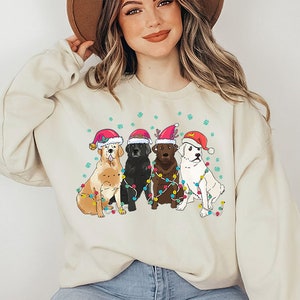 Puede incluir: Una sudadera de color crema con un dise&ntilde;o navide&ntilde;o que presenta cuatro perros con gorros de Pap&aacute; Noel y rodeados de luces de colores.