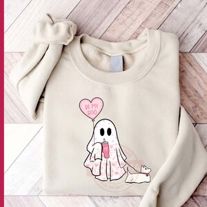 Puede incluir: Una sudadera blanca con un fantasma rosa que sostiene una bebida rosa y un globo de coraz&oacute;n rosa que dice "Be My Boo". El fantasma lleva un patr&oacute;n de coraz&oacute;n rosa y tiene un perro rosa con una correa.
