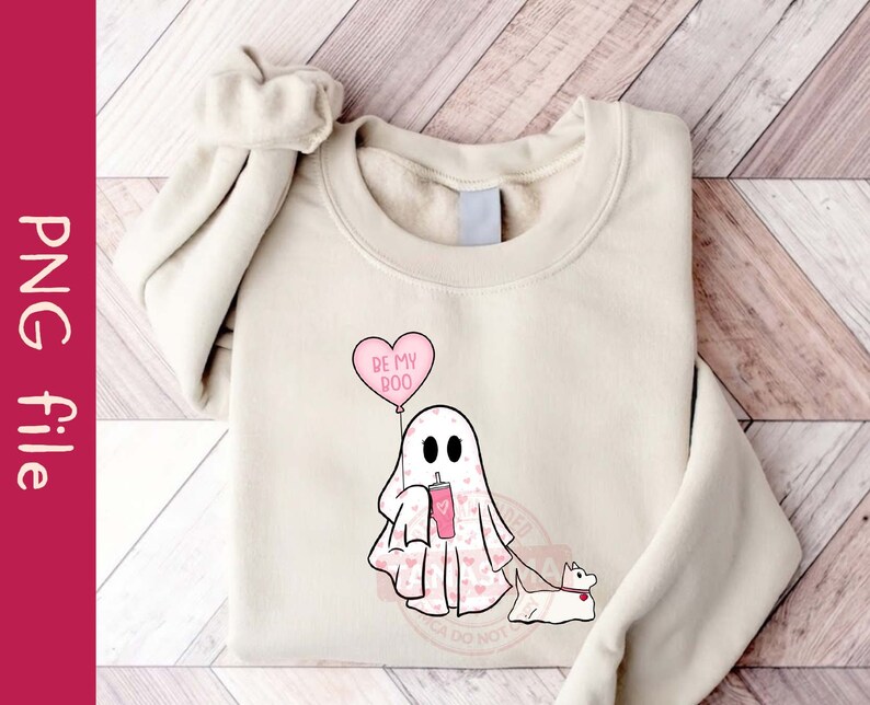 Puede incluir: Una sudadera blanca con un fantasma rosa que sostiene una bebida rosa y un globo de coraz&oacute;n rosa que dice "Be My Boo". El fantasma lleva un patr&oacute;n de coraz&oacute;n rosa y tiene un perro rosa con una correa.
